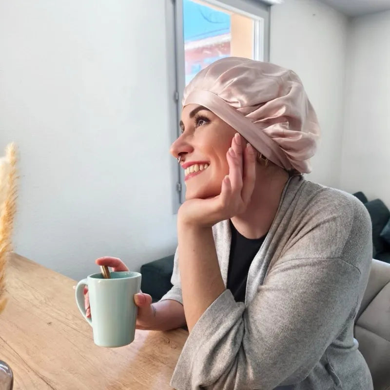 Femme souriante portant un bonnet en soie rose, tasse de café à la main – routine capillaire protectrice